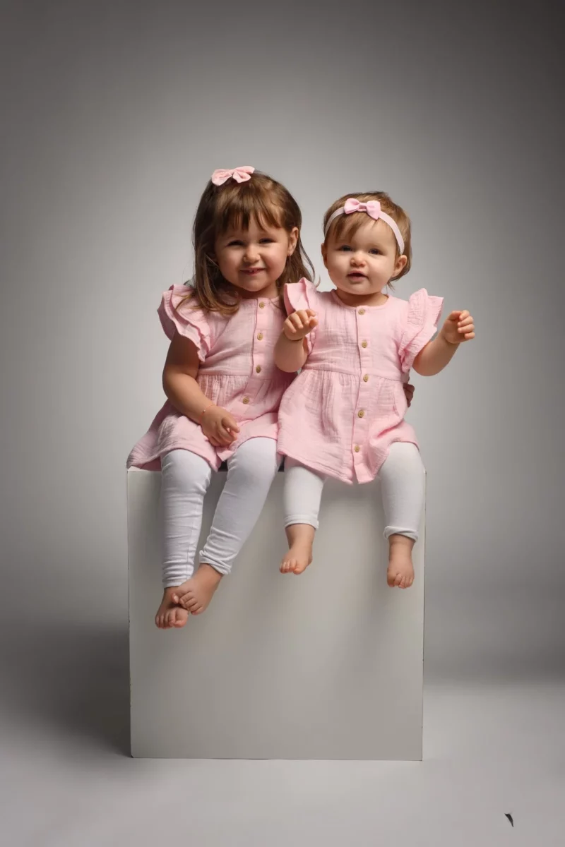 Deux jeunes filles sont assises sur un bloc blanc devant un fond neutre lors dune séance de photographie familiale en Seine-et-Marne. Toutes deux portent des robes roses assorties avec des leggings blancs. La fille la plus âgée sourit tandis que la plus jeune regarde devant elle, toutes deux avec des nœuds roses dans les cheveux.