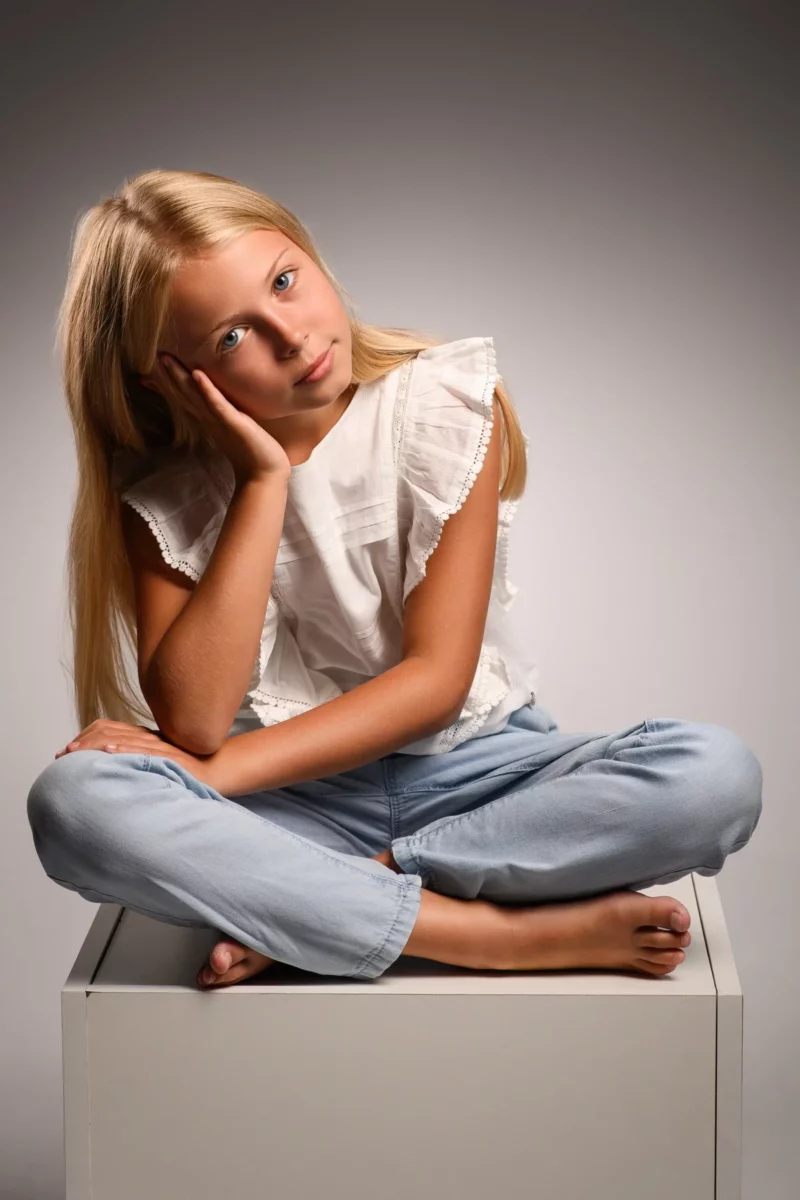Une jeune fille aux longs cheveux blonds est assise en tailleur sur un cube blanc. Elle porte un chemisier blanc à volants et un jean, la tête posée sur sa main. Sur fond gris, elle regarde directement lobjectif avec une expression détendue, capturant lessence dune séance de photographie en Seine-et-Marne.