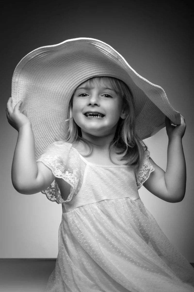 Une photo en noir et blanc montre une jeune fille souriante vêtue dune robe blanche et dun grand chapeau de soleil, tenant le bord à deux mains lors dune joyeuse séance de famille en Seine-et-Marne.