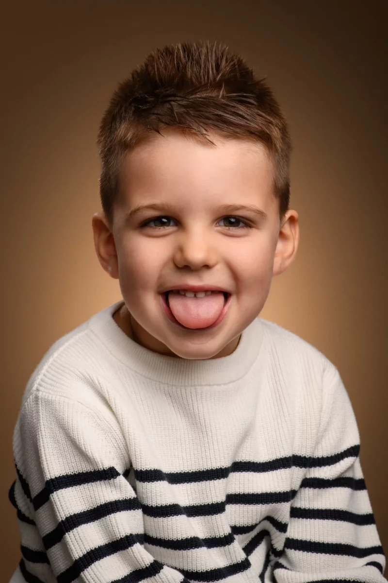 Un jeune garçon aux cheveux bruns courts tire la langue de manière enjouée lors dune séance photo de famille. Il porte un pull blanc à rayures noires et est assis sur un fond brun et chaud en Seine-et-Marne, souriant effrontément à lobjectif.