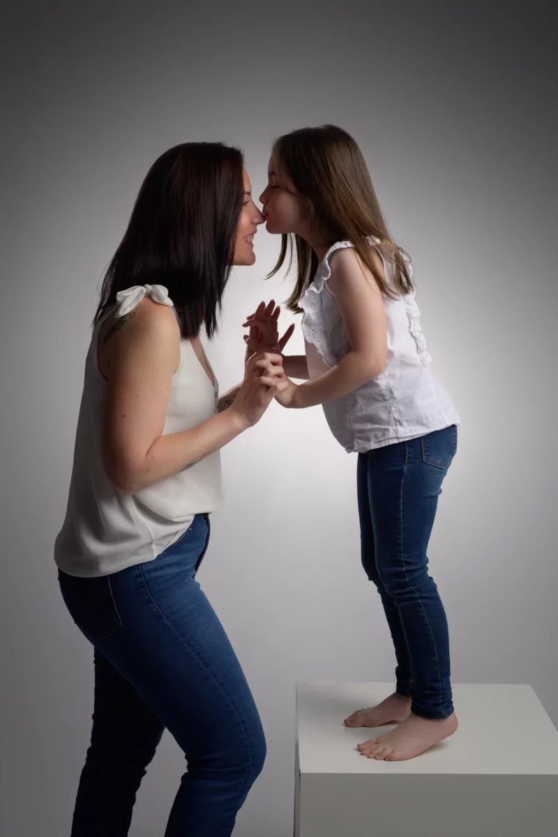 Une femme en haut blanc partage un moment de joie lors dune séance de photographie familiale, en se tenant la main et en souriant, recevant un baiser sur le nez dune jeune fille debout sur un bloc blanc. Toutes deux portent un jean bleu et ont de longs cheveux noirs, sur un fond gris rappelant le charme de la Seine-et-Marne.