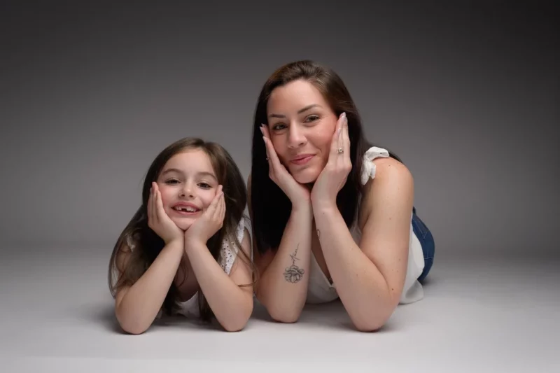Une femme et une jeune fille sont allongées sur le ventre, souriantes, le visage dans les mains, sur un fond gris. La femme, qui arbore un tatouage sur son poignet gauche, et la jeune fille sont vêtues de hauts blancs décontractés, capturant un moment de famille parfait lors de cette séance photo en Seine-et-Marne.