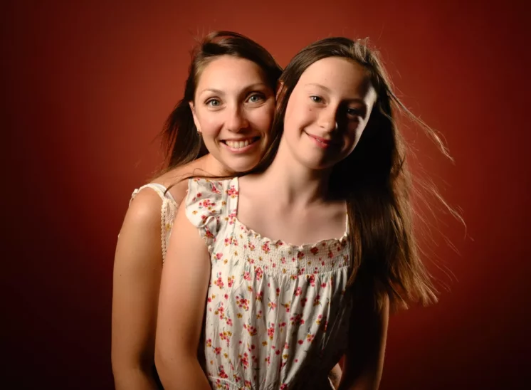 Une femme souriante et une jeune fille aux cheveux longs se tiennent devant un fond rouge chaleureux, magnifiquement capturées lors dune séance de photographie familiale en Seine-et-Marne. Toutes deux portent des hauts fleuris légers et sans manches tandis que la femme embrasse la jeune fille par derrière.