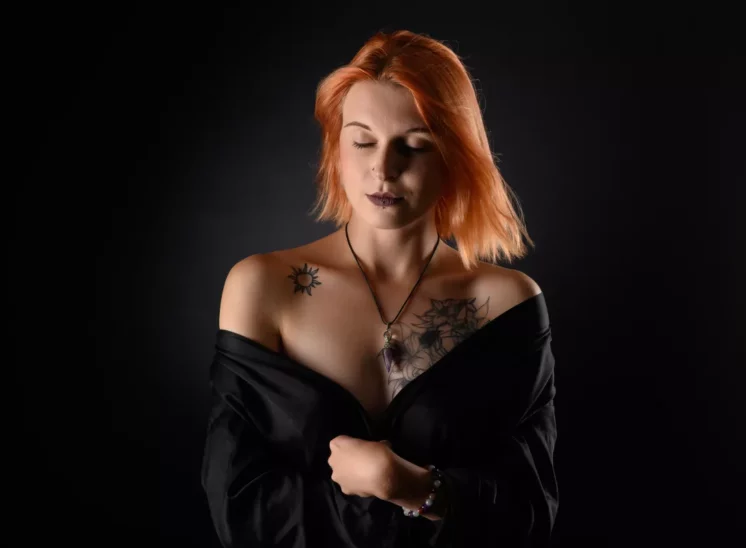 Une femme aux cheveux roux mi-longs pose devant un fond sombre, capturant un moment intime lors dune séance photo. Elle porte un haut noir à épaules dénudées, révélant des tatouages dun soleil et des motifs géométriques sur sa poitrine et son épaule. Ses yeux sont fermés, ornés dun collier et dun bracelet.