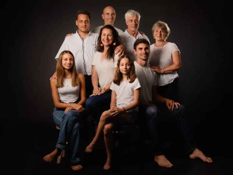 Une séance photo de groupe familiale en Seine-et-Marne capture trois générations, dont deux adultes plus âgés, trois adultes dâge moyen et deux jeunes filles. Vêtus de manière décontractée de hauts blancs et de jeans sur fond sombre, ils sourient chaleureusement à lobjectif.