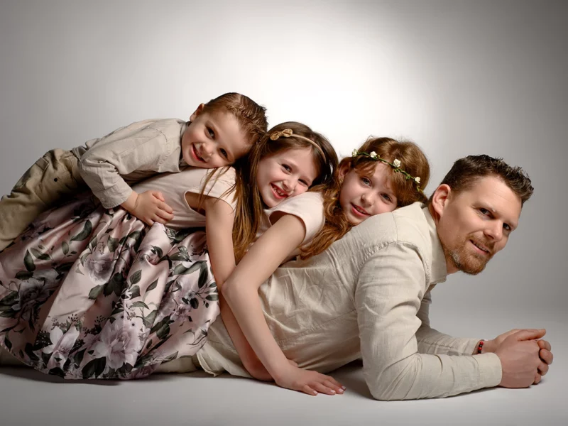 Un homme et trois enfants sont allongés les uns sur les autres, souriant chaleureusement. Les enfants, deux filles et un garçon, portent des tenues fleuries tandis que lhomme porte une chemise légère. Cette scène réconfortante capture lessence dune séance de en famille dans un décor neutre en Seine-et-Marne.