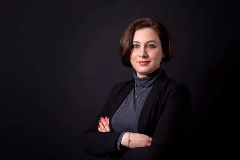 Une femme aux cheveux bruns courts se tient debout sur un fond sombre, incarnant lessence de lélégance dentreprise. Vêtue dun blazer noir et dun col roulé gris, ses bras sont croisés avec assurance avec du vernis à ongles rouge, parfaitement capturé par un photographe qualifié en Seine-et-Marne.