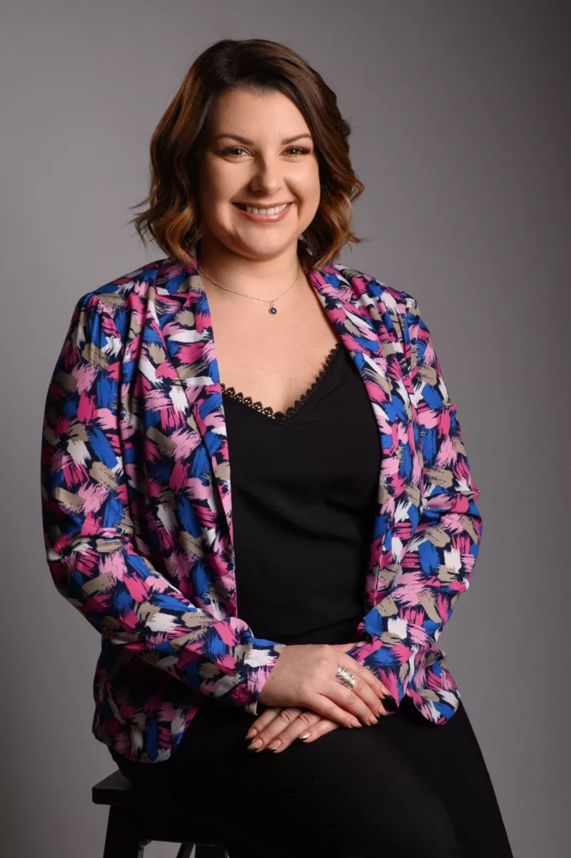 Une personne aux cheveux mi-longs sourit assise sur un tabouret, incarnant lessence de lélégance dentreprise. Elle porte un blazer fleuri coloré sur un haut noir sur fond uni. Photographiée en Seine-et-Marne, ses mains reposent gracieusement sur ses genoux.