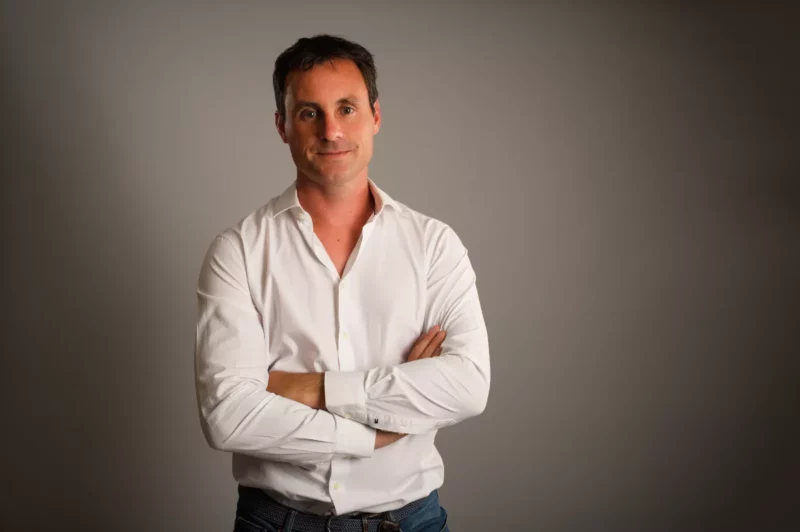 Un homme en chemise blanche et jean se tient debout, les bras croisés, souriant avec confiance sur un fond uni et sombre, illustrant le charme professionnel souvent capturé par les photographes dentreprise en Seine-et-Marne.