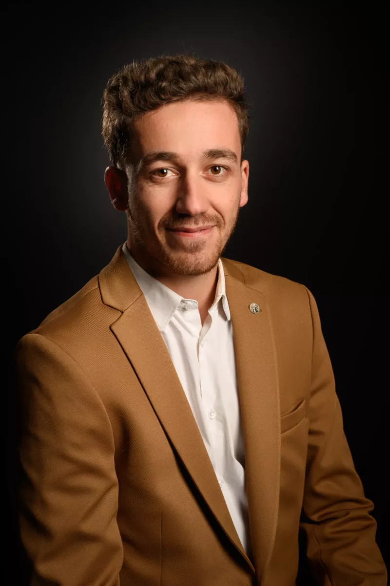 Un homme aux cheveux bruns courts et bouclés et à la barbe sourit. Vêtu dun blazer beige sur une chemise blanche, il respire la confiance. Le fond sombre renforce la chaleur de ce portrait professionnel, capturé par un photographe talentueux de Seine-et-Marne.