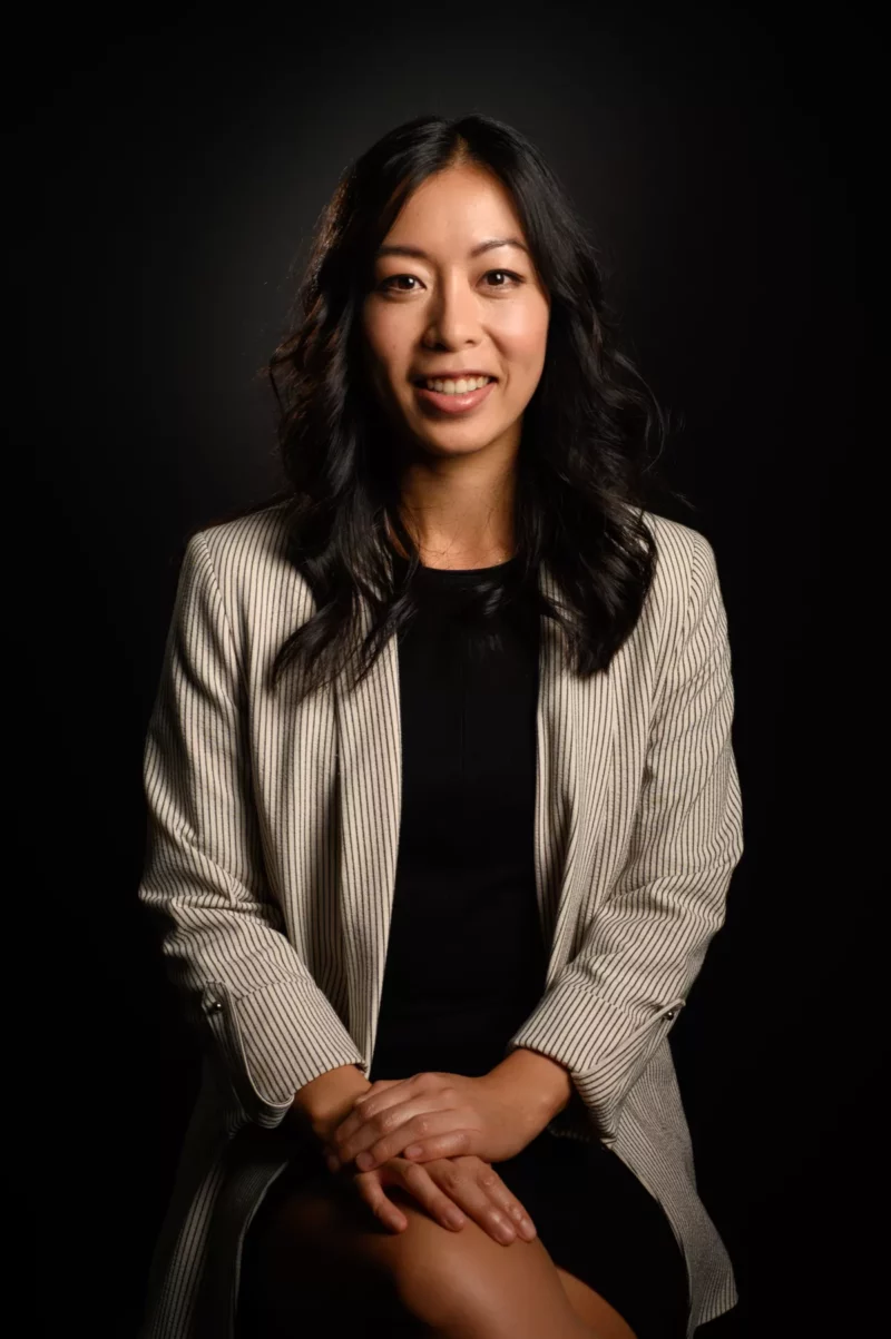 Dans un cadre dentreprise en Seine-et-Marne, une femme aux longs cheveux noirs est assise avec assurance sur un fond sombre, vêtue dun blazer à rayures claires sur un haut noir. Photographiée avec finesse, elle sourit doucement, les mains posées sur ses genoux.
