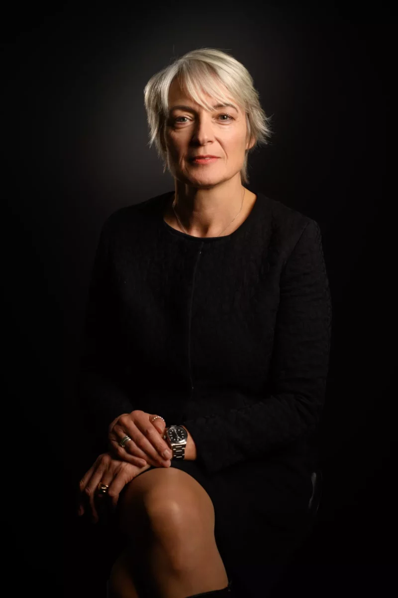 Une femme aux cheveux gris courts se tient assise avec assurance sur un fond sombre, incarnant lessence de la force dune entreprise. Vêtue dune tenue noire élégante et dune montre, elle présente une image parfaite pour lobjectif de tout photographe de Seine-et-Marne.