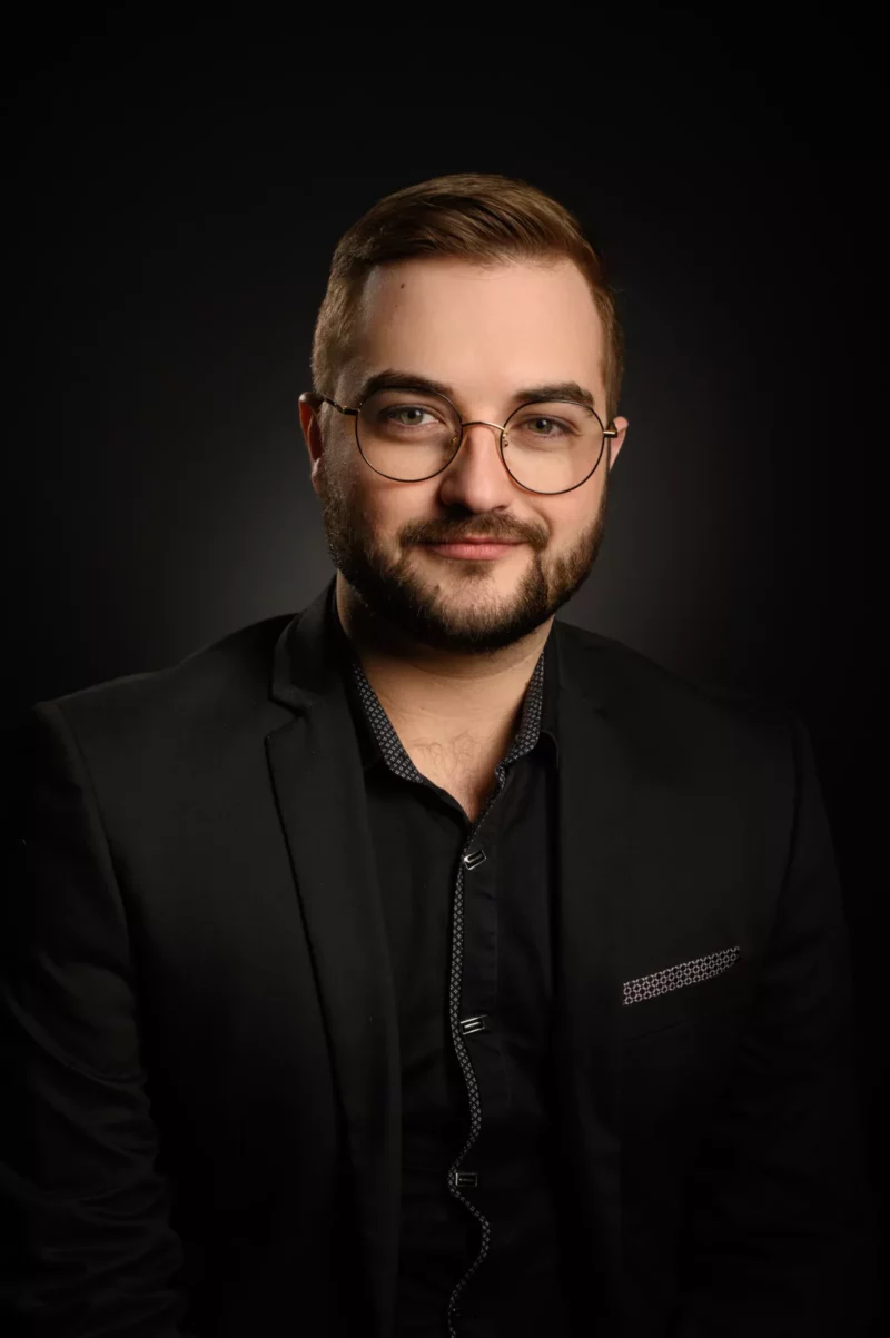 Un homme aux cheveux courts et à la barbe porte des lunettes et un costume noir avec une pochette. Il regarde lobjectif en souriant légèrement sur un fond sombre, capturant lessence de lélégance dentreprise dans ce portrait de Seine-et-Marne.
