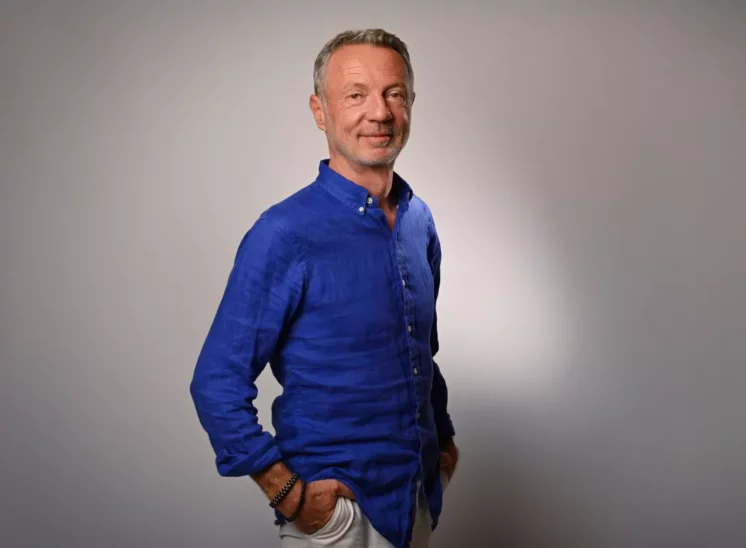 Un homme dâge moyen en chemise bleu vif se tient avec assurance sur un fond uni et clair, incarnant lessence de la sophistication dentreprise. Photographié avec une élégance subtile en Seine-et-Marne, il sourit chaleureusement, les mains dans les poches.