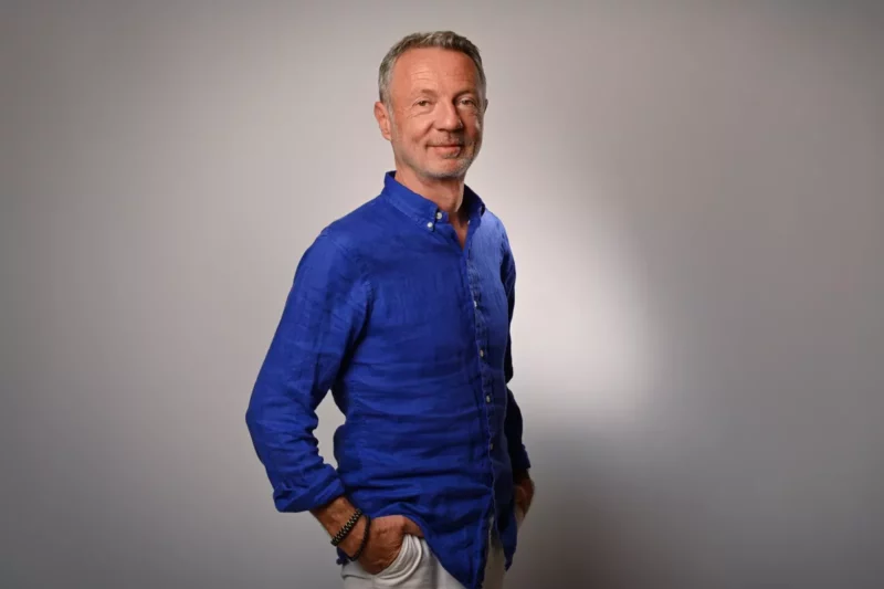 Un homme dâge moyen en chemise bleu vif se tient avec assurance sur un fond uni et clair, incarnant lessence de la sophistication dentreprise. Photographié avec une élégance subtile en Seine-et-Marne, il sourit chaleureusement, les mains dans les poches.