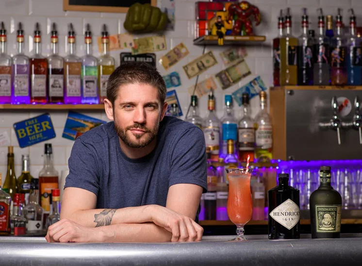 Un homme barbu est accoudé au comptoir dun bar de Seine-et-Marne, entouré de plusieurs bouteilles dalcool. Devant lui, un cocktail dans un grand verre et deux bouteilles de gin Hendricks, capturant lessence avec un style photographique corporate au milieu dune décoration colorée et éclectique.