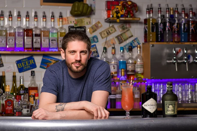 Un homme barbu est accoudé au comptoir dun bar de Seine-et-Marne, entouré de plusieurs bouteilles dalcool. Devant lui, un cocktail dans un grand verre et deux bouteilles de gin Hendricks, capturant lessence avec un style photographique corporate au milieu dune décoration colorée et éclectique.