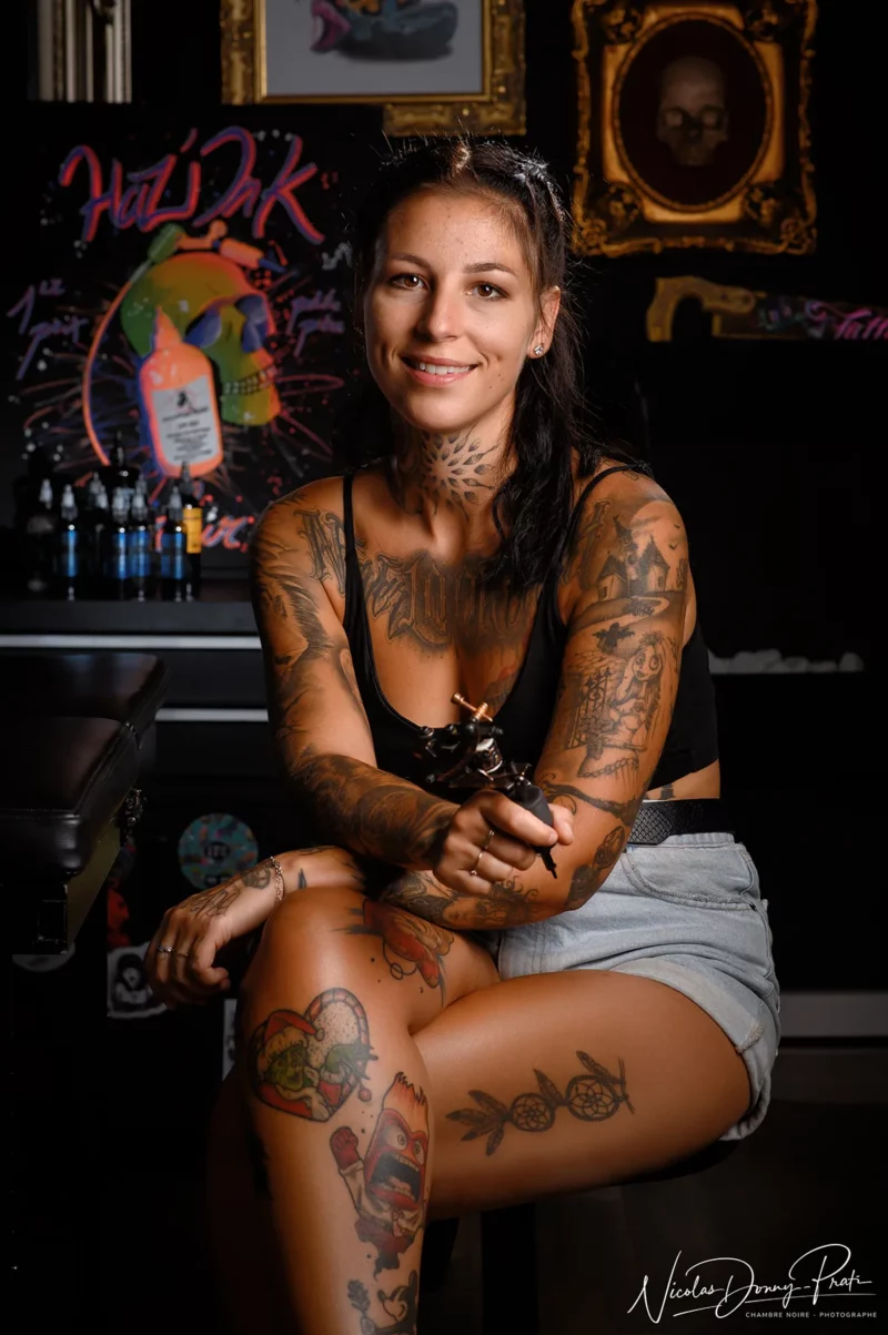 Une femme tatouée est assise avec assurance sur un tabouret dans un studio de tatouage en Seine-et-Marne, tenant une machine à tatouer. Elle porte un débardeur noir et un short en jean. Larrière-plan présente du matériel de tatouage coloré et des illustrations, magnifiquement capturées par un photographe dentreprise qualifié. Elle sourit à lobjectif.