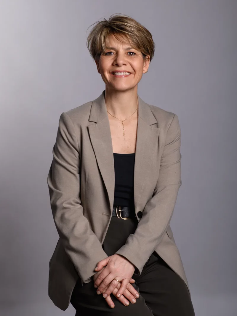 Une femme aux cheveux courts est assise sur un fond gris uni, incarnant lessence de lélégance dentreprise. Elle porte un blazer beige sur un haut noir et un pantalon foncé, les mains croisées sur ses genoux et un sourire chaleureux - un sujet parfait pour une photographie dentreprise en Seine-et-Marne.