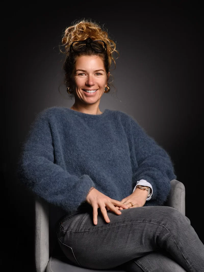 Une personne aux cheveux bouclés est assise sur une chaise, souriante, comme prête pour une séance photo dentreprise en Seine-et-Marne. Elle porte un pull bleu duveteux et un jean, les mains posées sur ses genoux. Le fond sombre et léclairage stratégique mettent magnifiquement en valeur son visage.
.