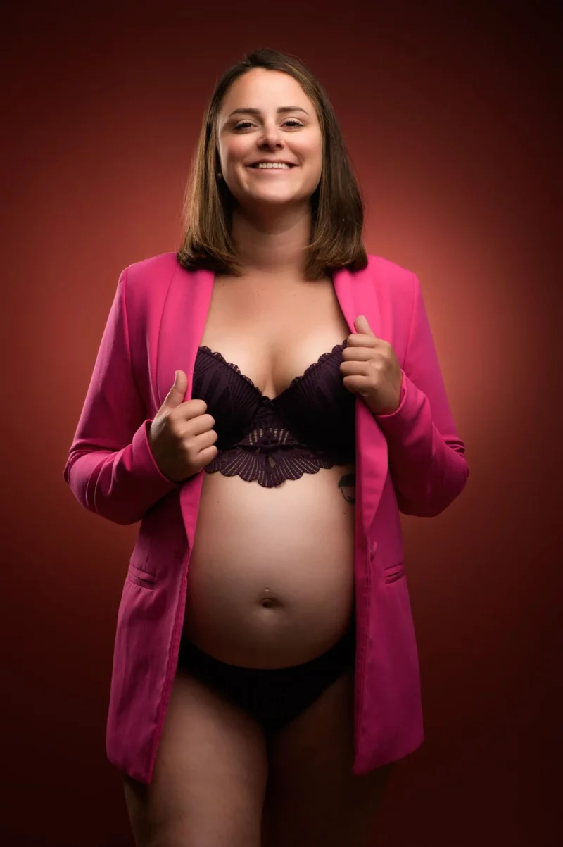 Une femme enceinte sourit tout en tenant les revers dun blazer rose vif, mettant en valeur sa beauté dans cette séance de Photographe Grossesse Seine-et-Marne (77). Elle porte un soutien-gorge et des sous-vêtements noirs sur un fond brun rougeâtre chaleureux.