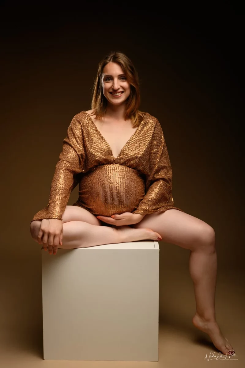 Une femme enceinte est assise sur un bloc blanc sur un fond brun chaleureux. Photographiée en Seine-et-Marne (77), elle porte une tenue dorée scintillante et sourit à lobjectif, les mains posées sur son ventre nu. Ses jambes sont détendues et écartées.