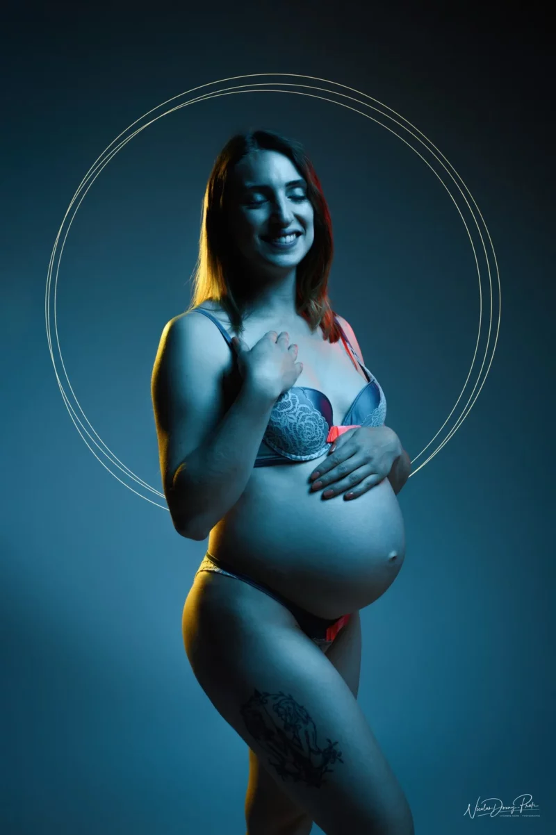 Une femme enceinte se tient debout, confiante, souriante, les yeux fermés et la main sur le ventre, magnifiquement capturée dans une séance de Photographe Grossesse Seine-et-Marne (77). Elle porte de la lingerie et exhibe un tatouage sur la cuisse, encadrée par une douce lumière circulaire sur fond sombre.