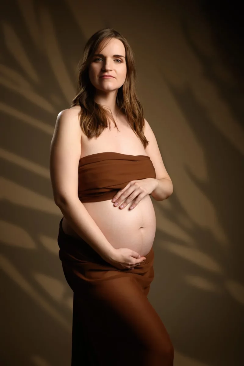 Une femme enceinte aux longs cheveux bruns se tient debout sur un fond marron, portant un foulard marron sans bretelles qui couvre sa poitrine et tombe. Elle berce doucement son ventre, regardant doucement lobjectif. Ce moment serein capture lessence du photographe grossesse Seine-et-Marne (77).