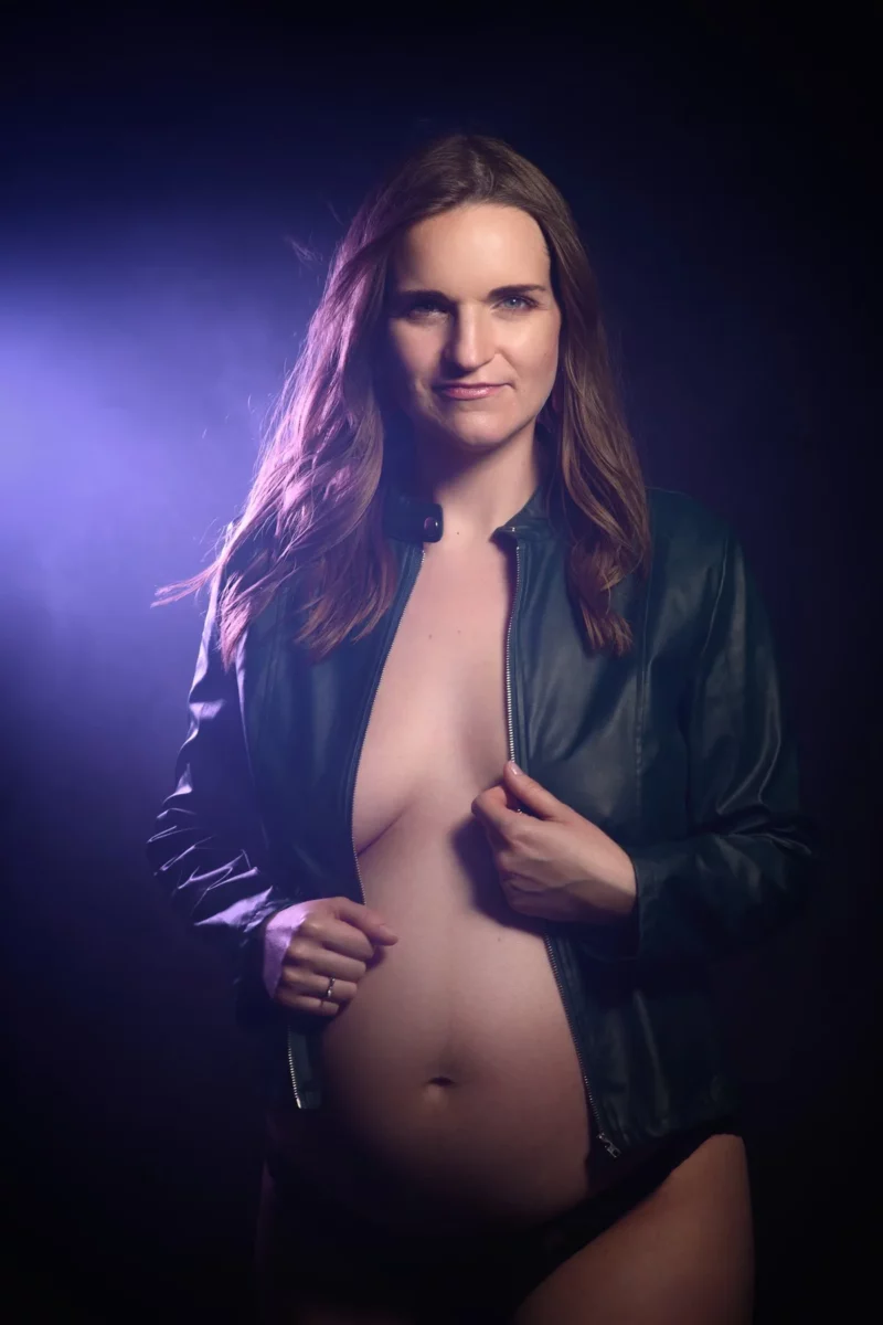 Une femme enceinte se tient debout, confiante, sur un fond sombre avec une lumière violette dans cette séance photo saisissante du Photographe Grossesse Seine-et-Marne (77). Elle porte une veste en cuir noir ouverte et un bas noir, révélant son ventre. Ses cheveux longs sont détachés et elle regarde directement lobjectif.