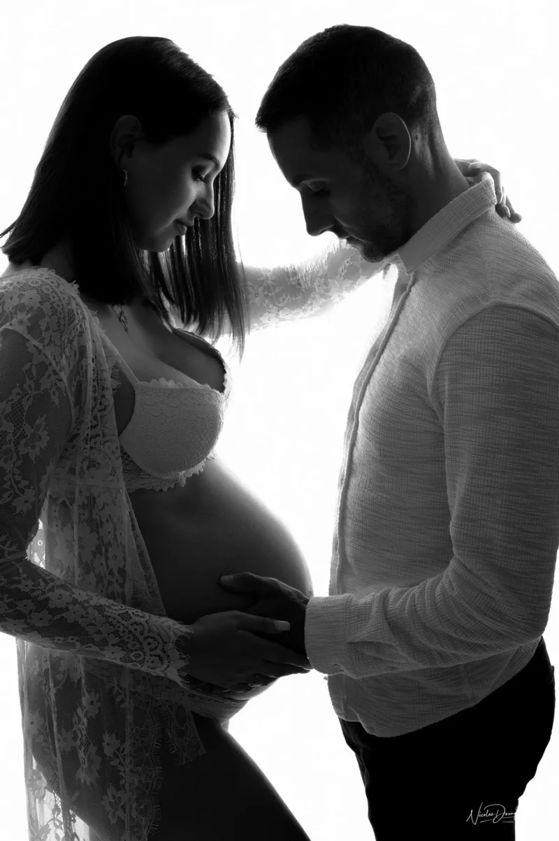 Silhouette dune femme enceinte et dun homme debout face à face. Capturée avec expertise par Photographe Grossesse Seine-et-Marne (77), la scène montre la femme en robe de dentelle et lingerie, tandis que lhomme, en chemise, pose doucement ses mains sur son ventre, incarnant lintimité et la tendresse.
