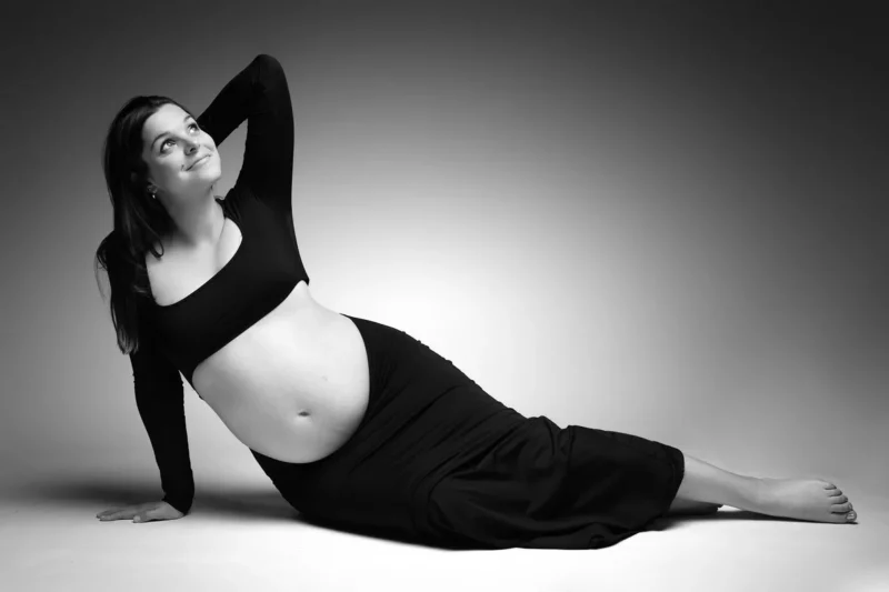 Photographie en noir et blanc dune femme enceinte en jupe longue et haut court. Elle est allongée sur le côté, la main droite soutenant sa tête et le bras gauche tendu derrière elle. Prise en photo par le photographe Grossesse Seine-et-Marne (77), elle regarde sereinement vers le haut sur un fond dégradé.