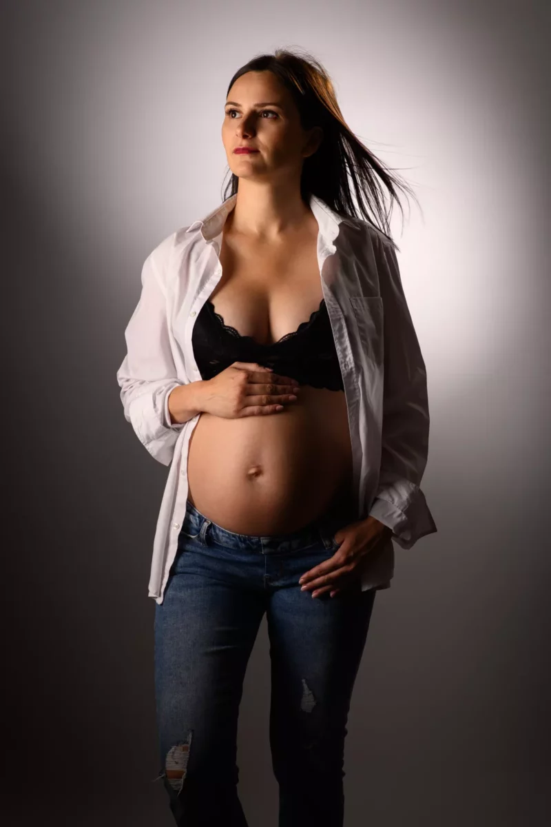 Une femme enceinte regarde vers le haut, vêtue dune chemise blanche partiellement ouverte, dun soutien-gorge noir et dun jean bleu. Ses cheveux sont doucement soufflés vers larrière alors quelle se tient debout, une main sur son ventre et lautre dans sa poche. Larrière-plan doucement éclairé capture lessence sereine dun photographe grossesse Seine-et-Marne (77).