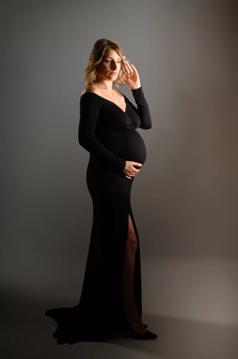 Une femme enceinte se tient gracieusement sur un fond gris dans une longue robe noire, incarnant lessence du photographe grossesse Seine-et-Marne (77). Une main repose doucement sur son ventre tandis que lautre touche ses cheveux, tandis quelle regarde pensivement sur le côté.