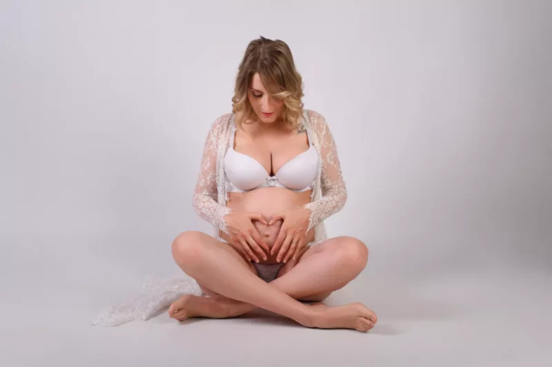 Dans un moment serein capturé par le photographe Grossesse Seine-et-Marne (77), une femme enceinte est assise en tailleur sur le sol, portant un soutien-gorge blanc et un cache-maillot en dentelle. Ses mains forment doucement un cœur sur son ventre, sur fond gris clair uni.