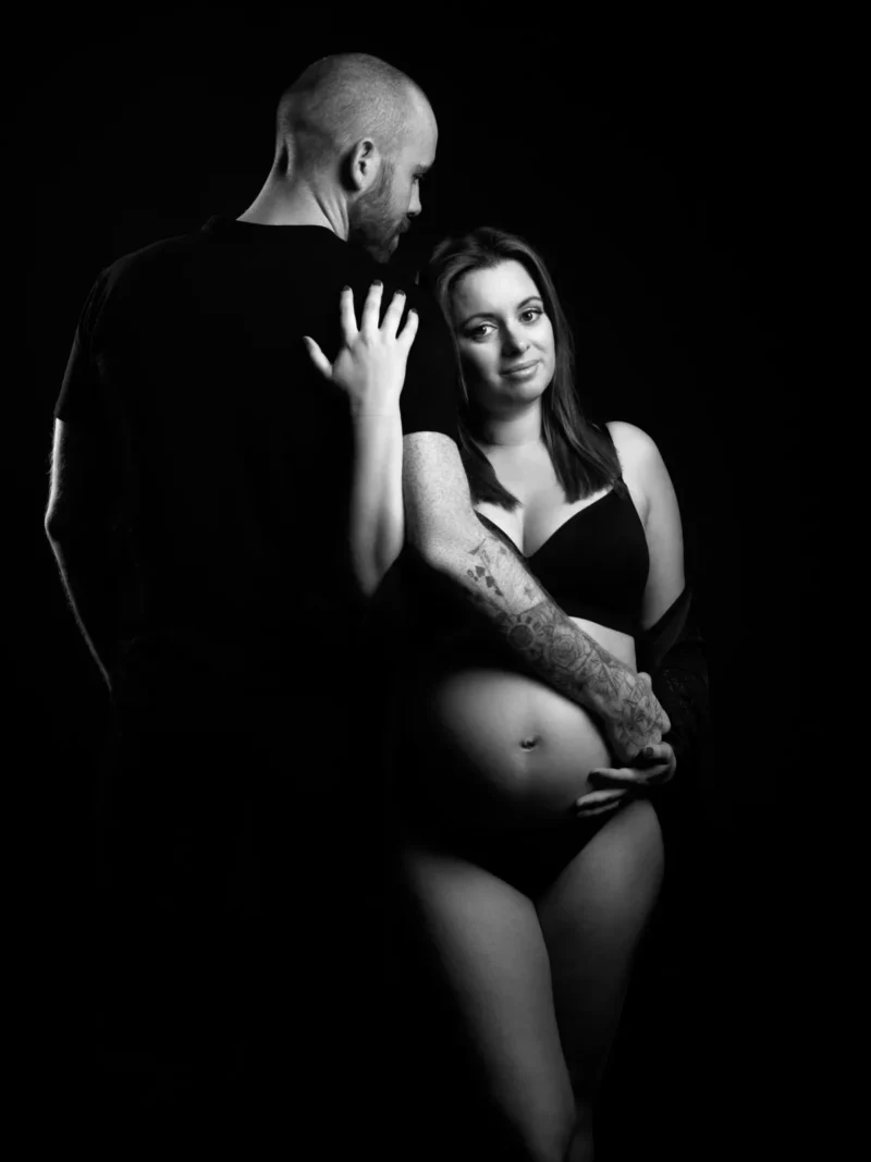 Une photo en noir et blanc montre une femme enceinte en soutien-gorge et sous-vêtements noirs, souriant tout en tenant son ventre. À côté delle se tient un homme en t-shirt noir, lenlaçant dun bras autour de ses épaules - un beau moment du photographe Grossesse Seine-et-Marne (77).