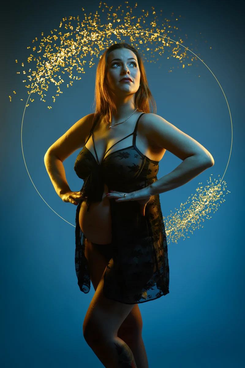 Une femme se tient debout, confiante, en lingerie sur un fond bleu, entourée dun halo de paillettes dorées. Elle regarde vers le haut, les mains sur les hanches, illuminée par une lumière chaude. Cette scène captivante pourrait facilement être présentée dans une vitrine de photographe grossesse Seine-et-Marne (77).