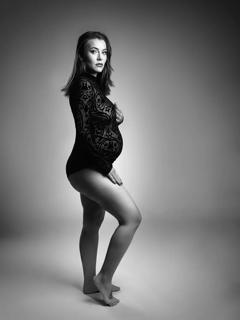 Photographie en noir et blanc dune femme enceinte debout, pieds nus, de profil, prise par un talentueux photographe grossesse en Seine-et-Marne (77). Elle porte une tenue moulante à motifs et touche son ventre avec une expression sereine sur un fond doucement éclairé.