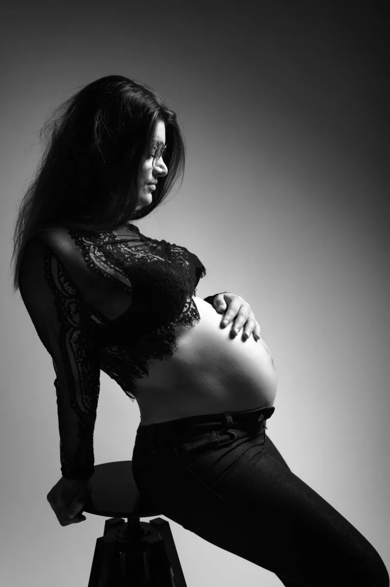 Photographie en noir et blanc prise par le photographe Grossesse Seine-et-Marne (77) montrant une femme enceinte assise sur un tabouret, vêtue dun haut en dentelle et dun jean foncé. Elle pose doucement ses mains sur son ventre et regarde sur le côté, éclairée par une douce lumière venant den haut.