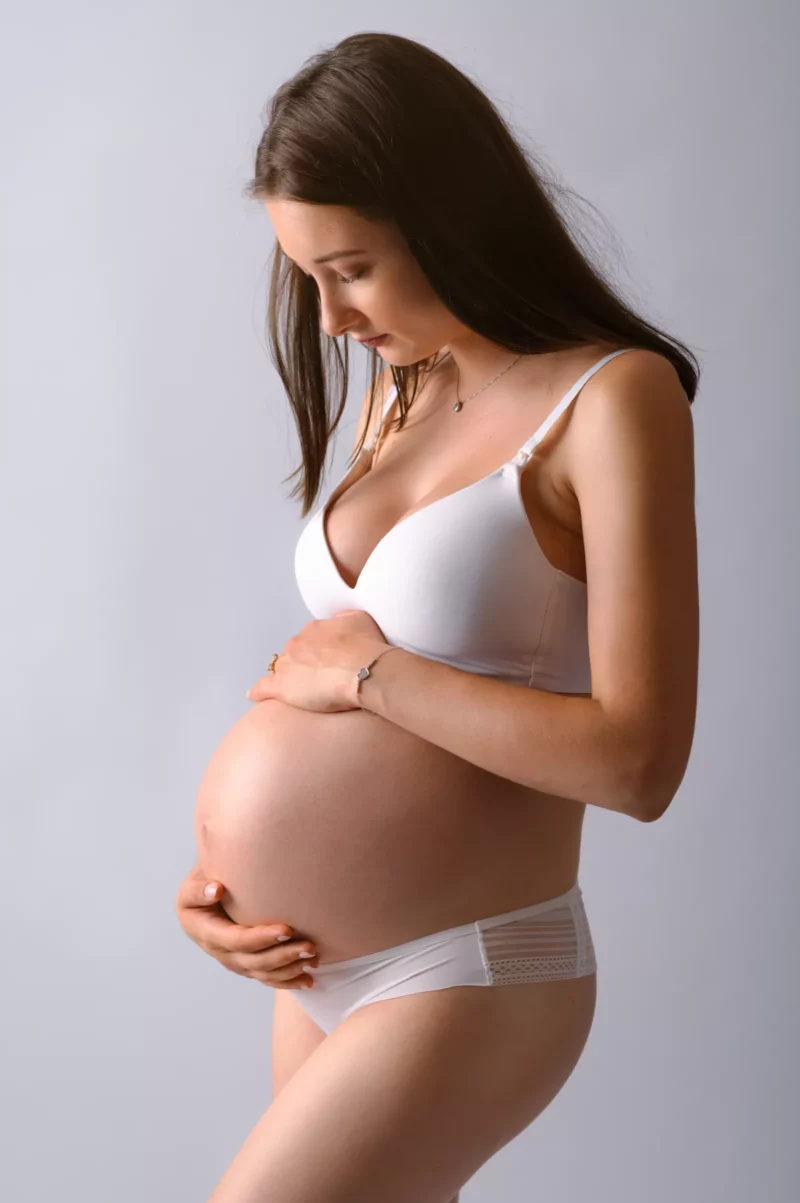 Une femme enceinte en lingerie blanche berce amoureusement son ventre rond, magnifiquement capturée par le photographe Grossesse Seine-et-Marne (77). Elle regarde vers le bas avec un doux sourire sur un fond uni et clair, transmettant un moment serein et intime de maternité.