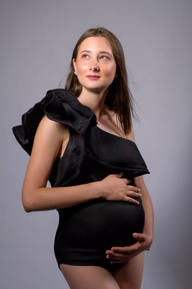 Une femme enceinte, vêtue dune tenue noire asymétrique, se tient gracieusement sur un fond gris. Elle tient son ventre à deux mains, reflétant le talent artistique du photographe Grossesse Seine-et-Marne (77), et regarde sur le côté avec une expression pensive.
