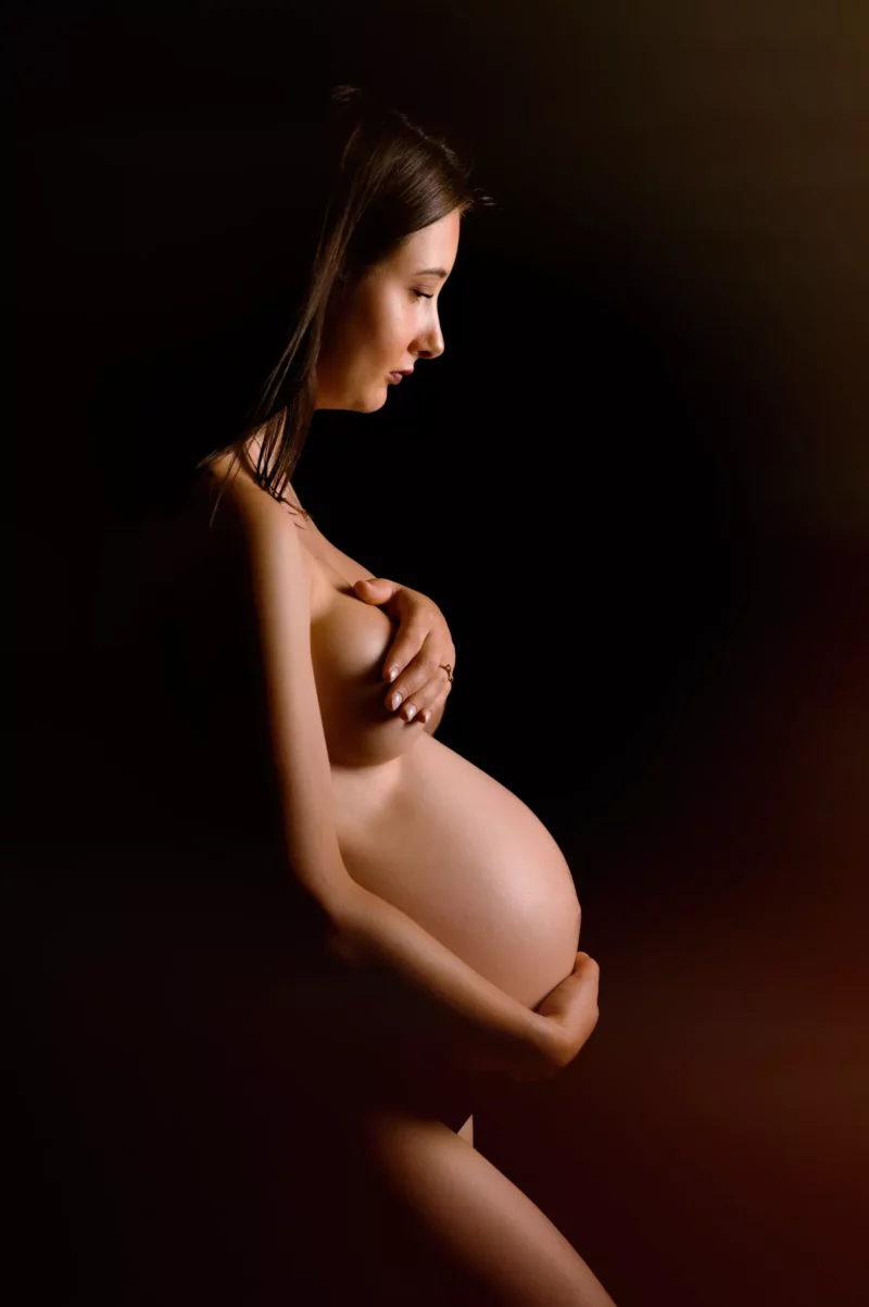 Profil dune femme enceinte tenant son ventre dune main et couvrant sa poitrine de lautre, capturée dans une séance exquise de Photographe Grossesse Seine-et-Marne (77). Elle se tient debout sur un fond sombre et doucement éclairé, le regard baissé avec une expression sereine.