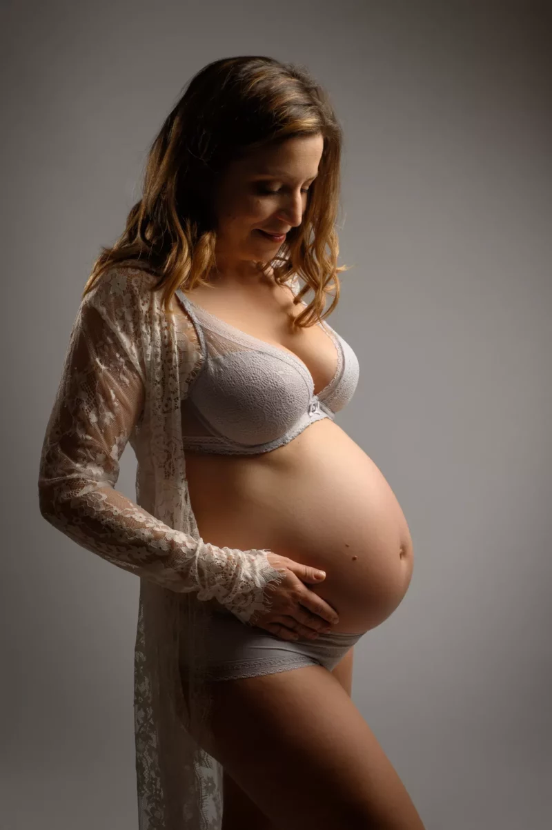 Une femme enceinte se tient debout sur un fond neutre, berçant doucement son ventre. Vêtue dune robe légère en dentelle sur un soutien-gorge et des sous-vêtements, son expression est sereine et tendre tandis quelle regarde vers le bas - un beau moment capturé par le photographe grossesse Seine-et-Marne (77).