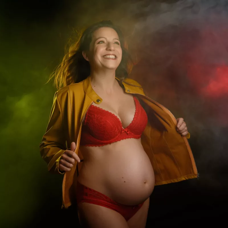 Une femme enceinte sourit avec assurance, vêtue de lingerie rouge et dune veste jaune moutarde. Elle est encadrée par un éclairage diffus coloré avec des nuances de vert et de rouge en arrière-plan, magnifiquement capturé par le photographe Grossesse Seine-et-Marne (77).