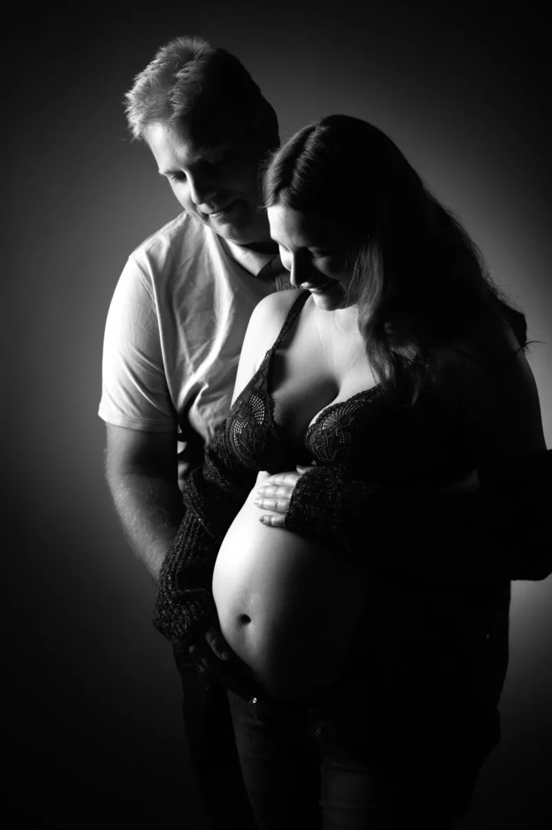 Une photo en noir et blanc montre une femme enceinte aux cheveux longs, en soutien-gorge et pull ouvert, tenant doucement son ventre. Un homme se tient derrière elle, la main sur son épaule, lui souriant dans ce décor à la lumière tamisée, illustrant parfaitement lessence du photographe grossesse Seine-et-Marne (77).