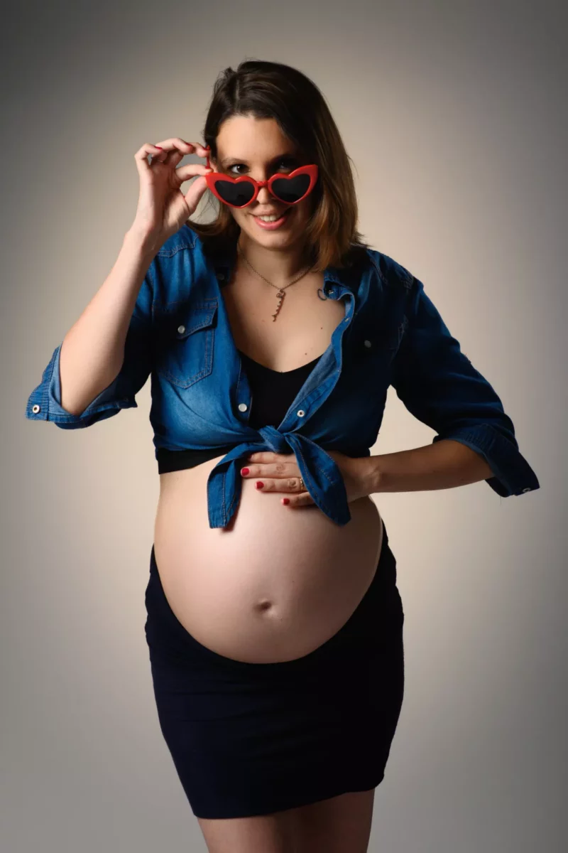 Une femme enceinte, photographiée dans une charmante séance de Photographe Grossesse Seine-et-Marne (77), porte une chemise en jean nouée et une jupe noire. Une main sur son ventre et lautre ajustant de manière ludique des lunettes de soleil rouges en forme de cœur, elle sourit à lobjectif sur un fond neutre.