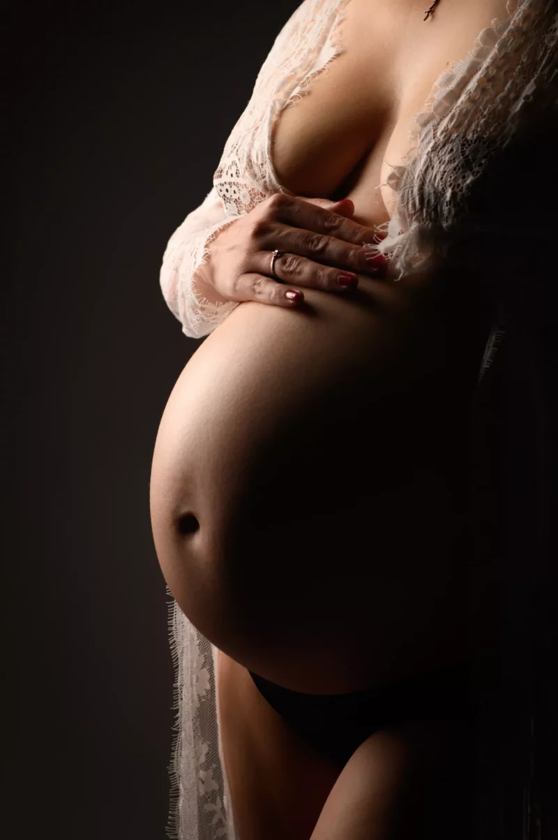 Une femme enceinte berce doucement son ventre, vêtue dun vêtement en dentelle drapé sur ses épaules. Prise dans un style de photographie de grossesse propre à la Seine-et-Marne (77), limage est doucement éclairée, soulignant la courbe du ventre sur le fond sombre.