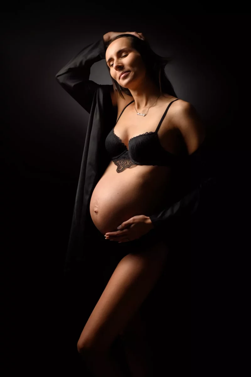 Une femme enceinte en lingerie noire pose sur un fond sombre, incarnant lart du photographe Grossesse Seine-et-Marne (77). Elle tient gracieusement son ventre tout en touchant ses cheveux, avec un éclairage capturant son expression sereine et sa forme intime.