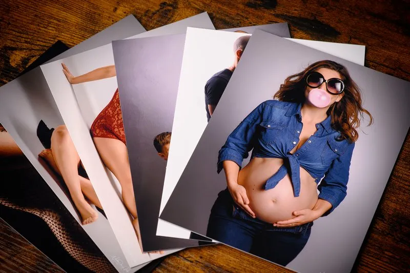 Une pile de photographies sur une surface en bois montre une femme en chemise en jean avec un ventre rond, soufflant une bulle de chewing-gum. Dautres images, prises par un photographe de Seine-et-Marne (77), la présentent dans diverses poses et tenues, notamment de la lingerie et des vêtements décontractés.