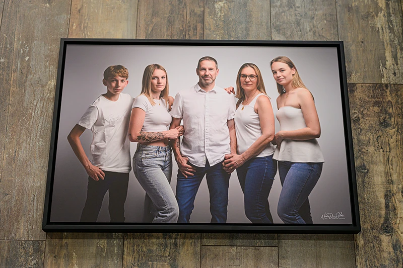 Un portrait de famille encadré, réalisé par un photographe de Seine-et-Marne (77), montre deux hommes et trois femmes vêtus de hauts blancs et de jeans bleus, se tenant ensemble sur un fond clair. La photo est joliment exposée sur un mur en bois.