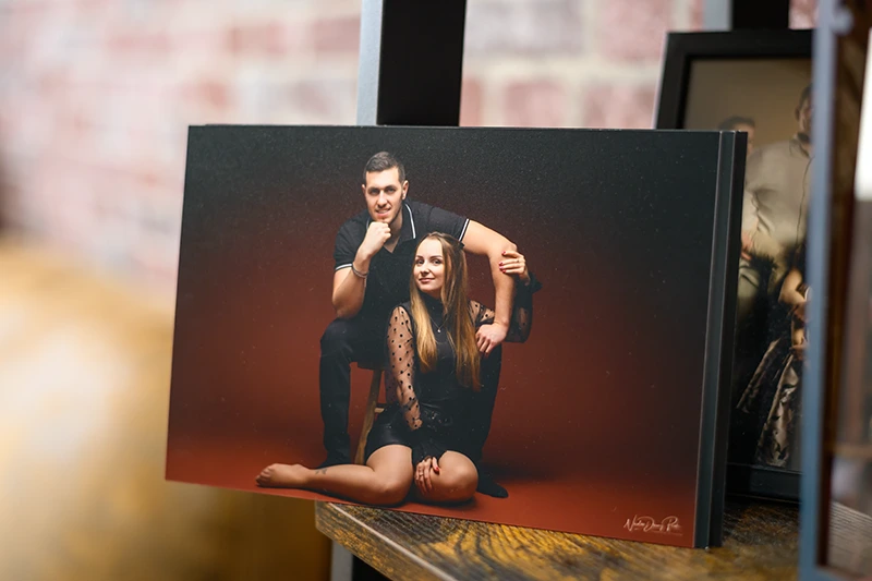 Une photo encadrée d'un photographe de Seine-et-Marne (77) montre un homme et une femme souriants, assis ensemble sur le sol, exposés sur une étagère en bois parmi d'autres photos encadrées chères à l'arrière-plan.